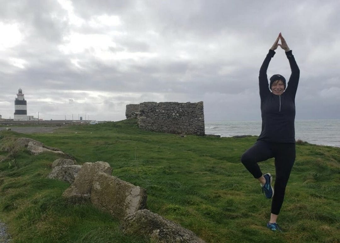 woman yogi in black top black pants running shoes in tree pose on grass in front of stone building and lighthouse in wickelow ireland