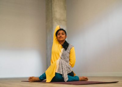 Svikami wearing yellow and black and white sari over blue yoga pants seated with left leg over right right arm over head grabbing left hand behind back eyes closed at My Vinyasa Practice studio in Austin Texas