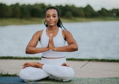 black woman yogi in white top and white pants sits cross legged hands pressed to heart's center on green mat in front of sidewalk in front of lake in a park