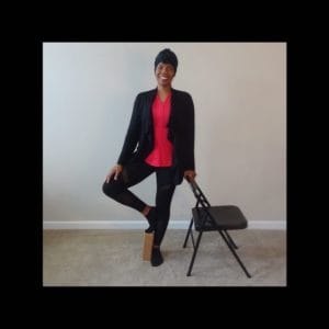 black woman yogi in black sweater red top smiling standing in tree pose left hand on folding metal chair right toes supported by cork block on carpet
