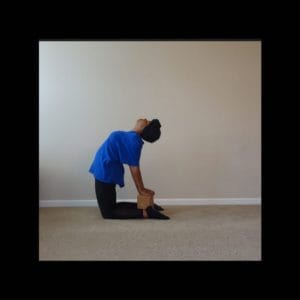 black woman yogi in blue top black bottoms kneels on carpet arms reach back to rest on cork block on back of ankles