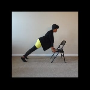 black woman yogi in black sweater yellow dress and black bottoms leans forward supported by folding metal chair on carpet for supported plank pose