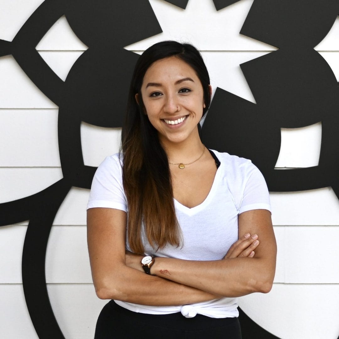Kristiana Vuong standing smiling wearing white top and black bottoms with arms crossed in front of the mandala at my vinyasa practice in austin texas