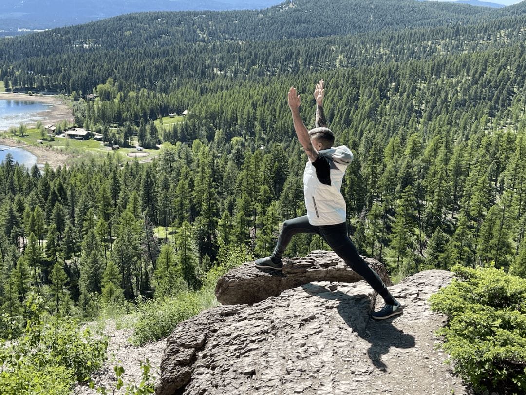A male yogi in white vest and black bottoms poses in crescent lunge pose on top of a boulder on the edge of a mountain with a mountain lake and pine forest behind him at Glacier National Park