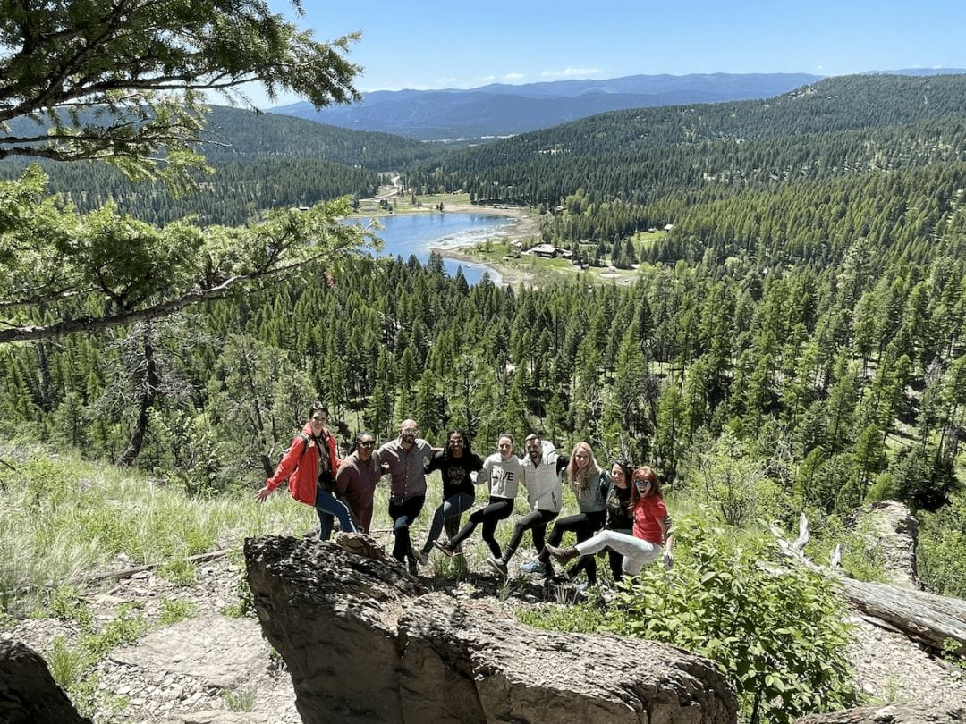 9 yogis posing on the edge of a cliff with a mountain lake and pine forest behind them under a blue sky in glacier national park