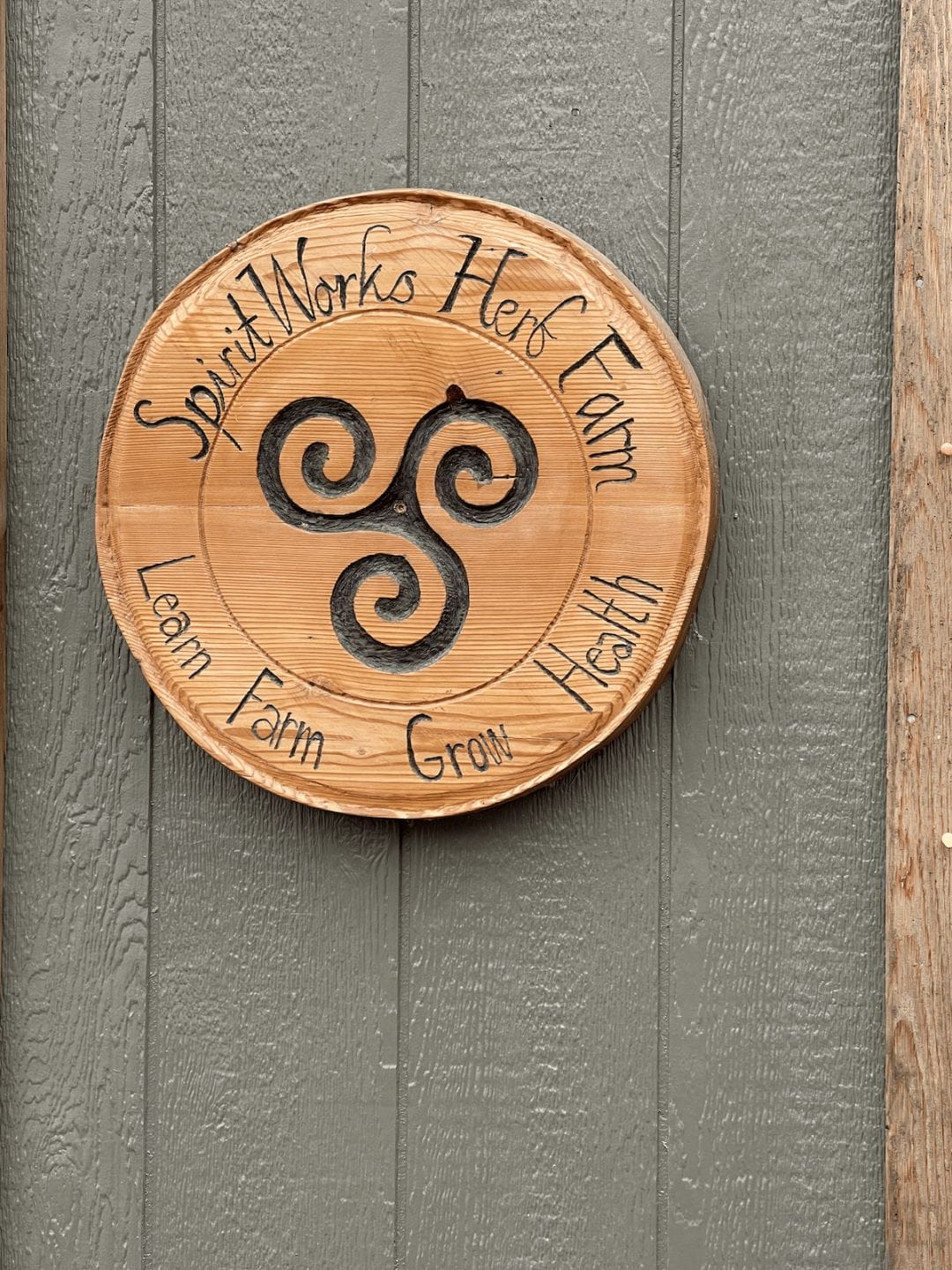 photo of a round wood plaque on the side of a grey exterior wall with text that reads "spirit works herb farm, learn farm grow health"
