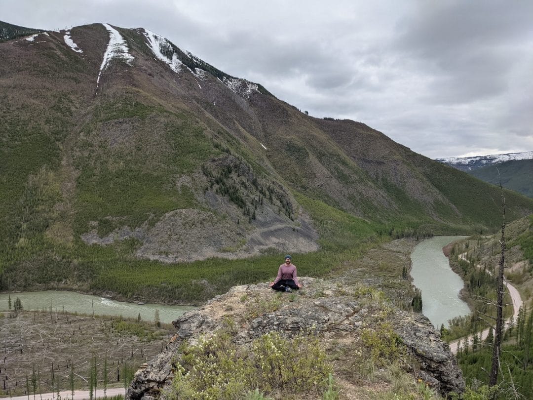 a male yogi in purple long sleeve top and black bottoms sits in lotus pose with eyes closed on a boulder with a green river and snow covered hills in the background at Glacier National Park