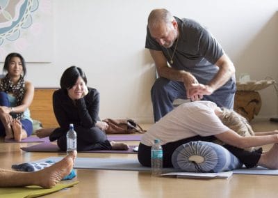 Leslie Kaminoff in grey t-shirt gold chain and grey pants kneels above a yogi in white top and black bottoms on a blue yoga mat in forward fold pose in front of 3 other yogis watching in a yoga studio