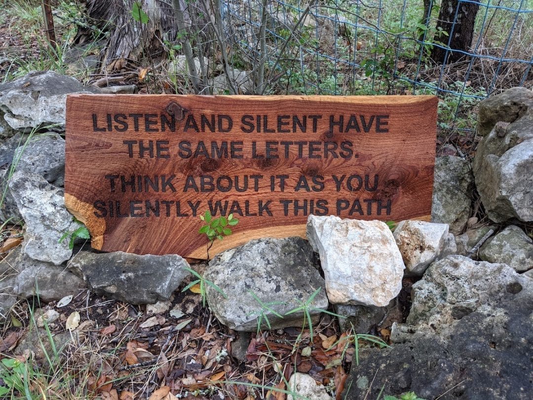 wood sign on the ground supported by rocks at the beginning of a garden labyrinth in north carolina with text that reads "listed and slient have the same letters, think about it as you silently walk this path"