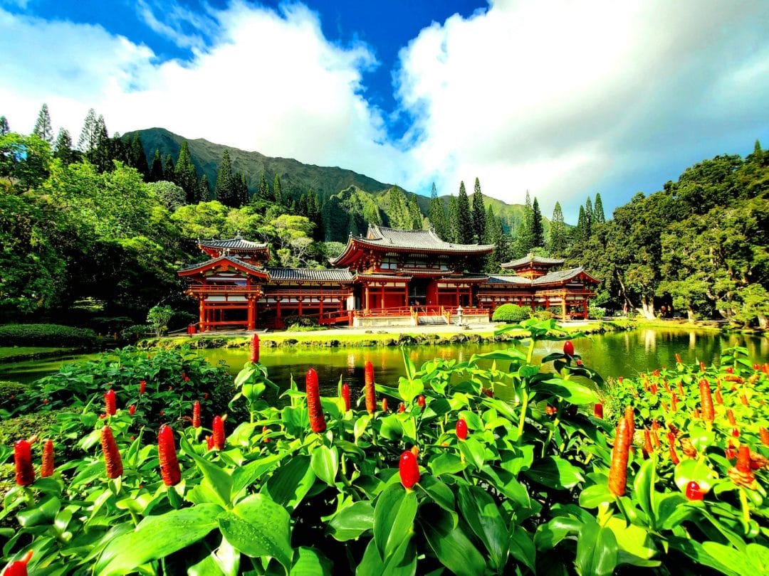 a japanese temple sits on a small island surrounded by water in a lush jungle with volcanic green hills in the background under a blue and partly cloudy sky