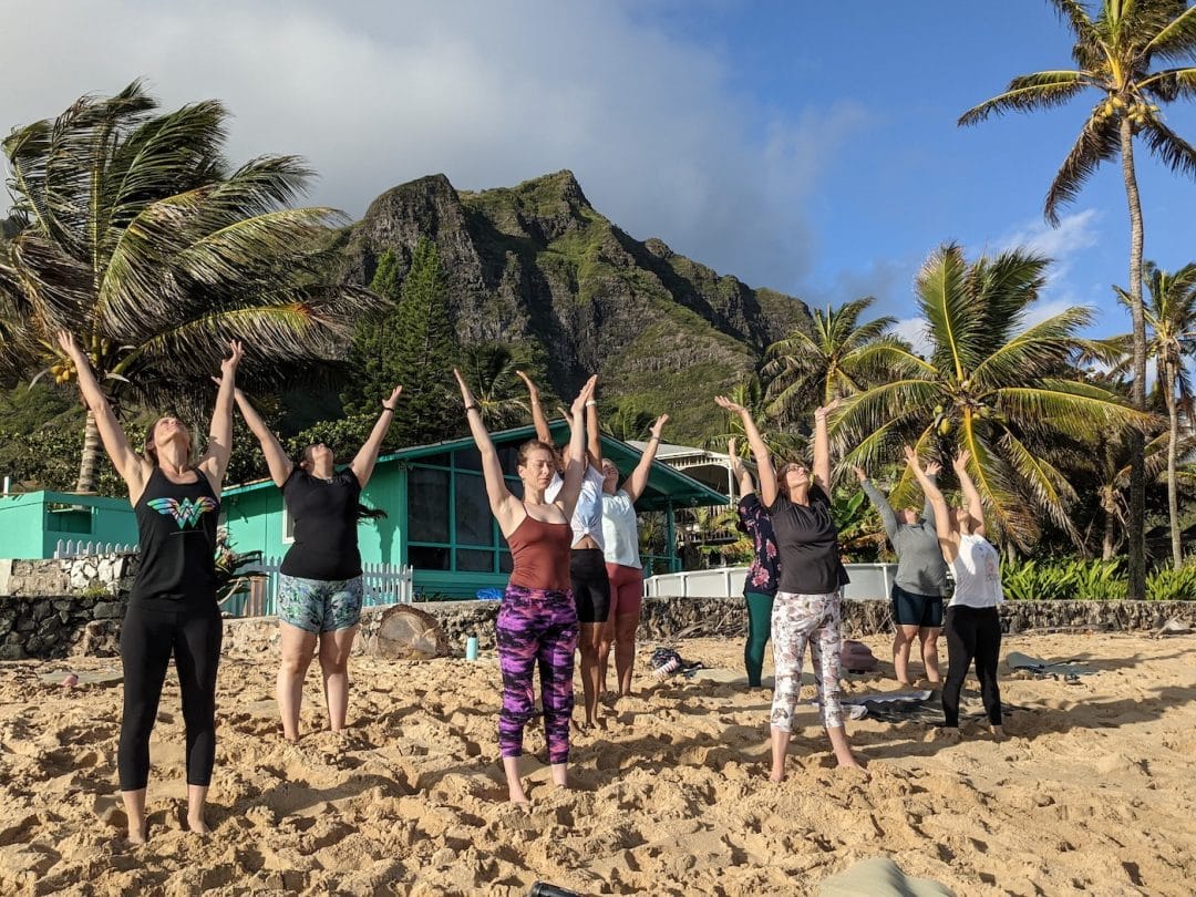 8 women yogis and one male yogi pose in a sun salutation on the beach in Hawaii with green volcanic mountains and partly cloudy blue sky in the background