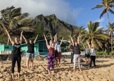 8 women yogis and one male yogi pose in a sun salutation on the beach in Hawaii with green volcanic mountains and partly cloudy blue sky in the background