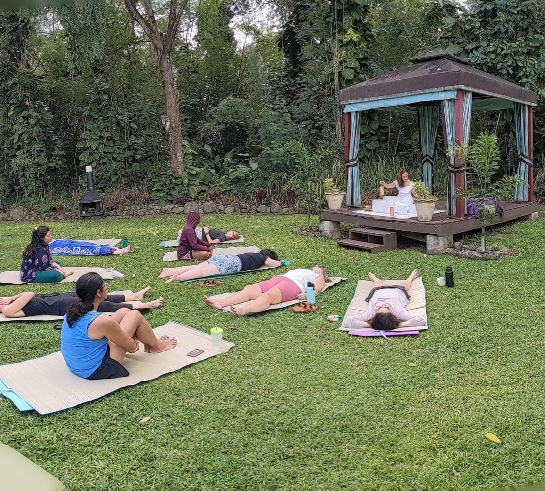 9 women yogis lay and sit on grass mats and yoga mats in a lush yard in front of a woman in a white dress on a raised gazebo who is performing a ceremony