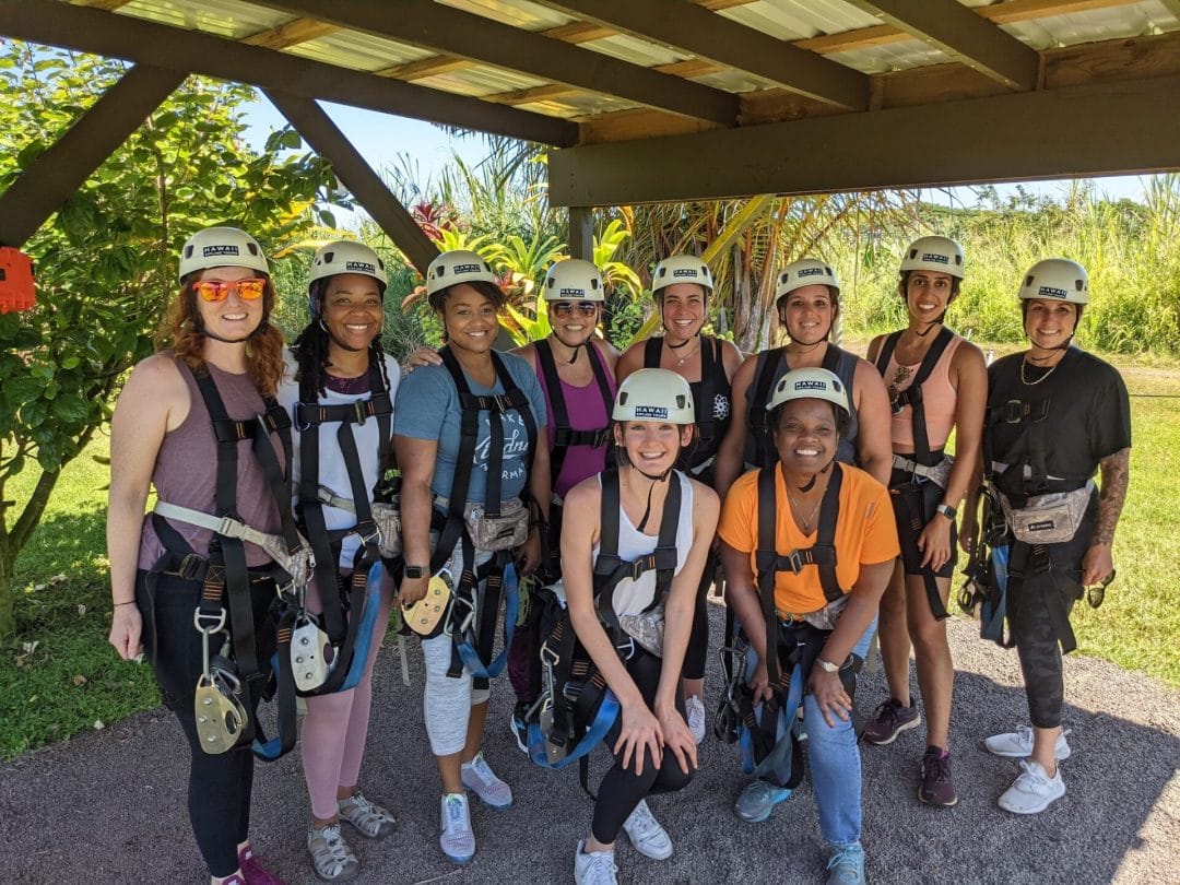 Michelle Young, Tara Cleven, and 8 women yogis in ziplining equipment posing under a shade structure at a park in Hawaii