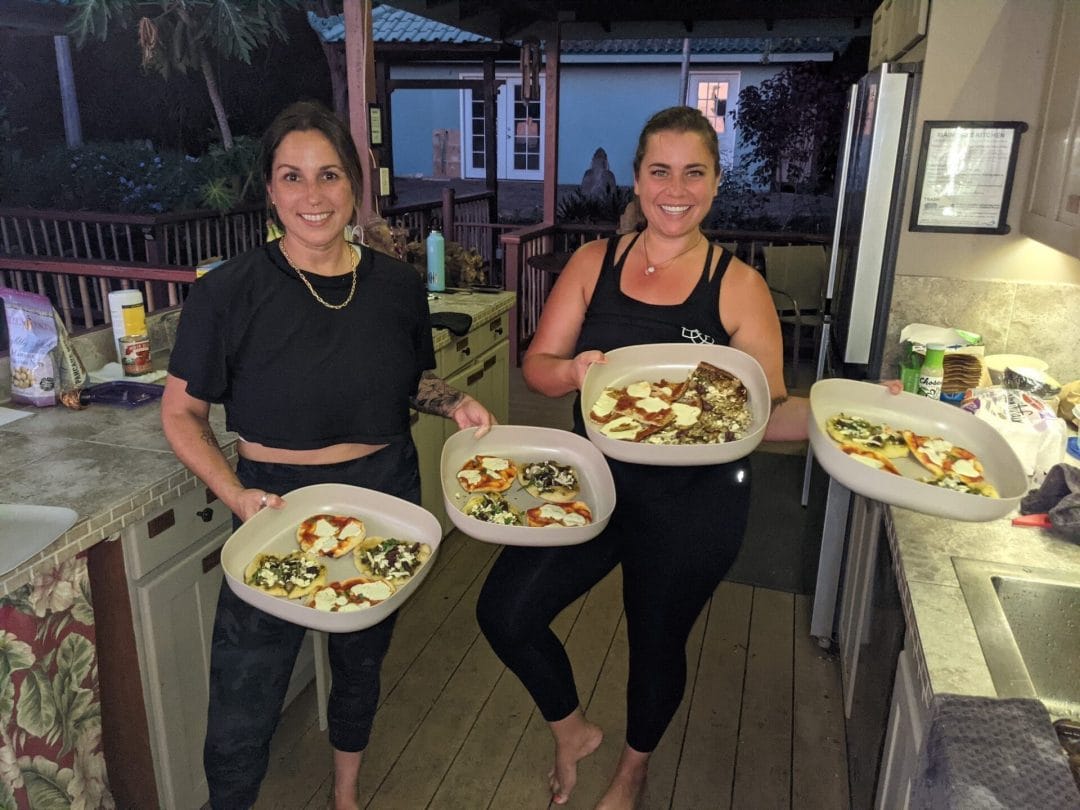 Michelle Young and Tara Cleven hold 4 earthen dishes with homemade pesto and margharita pizzas in an outdoor kitchen in a Hawaii house