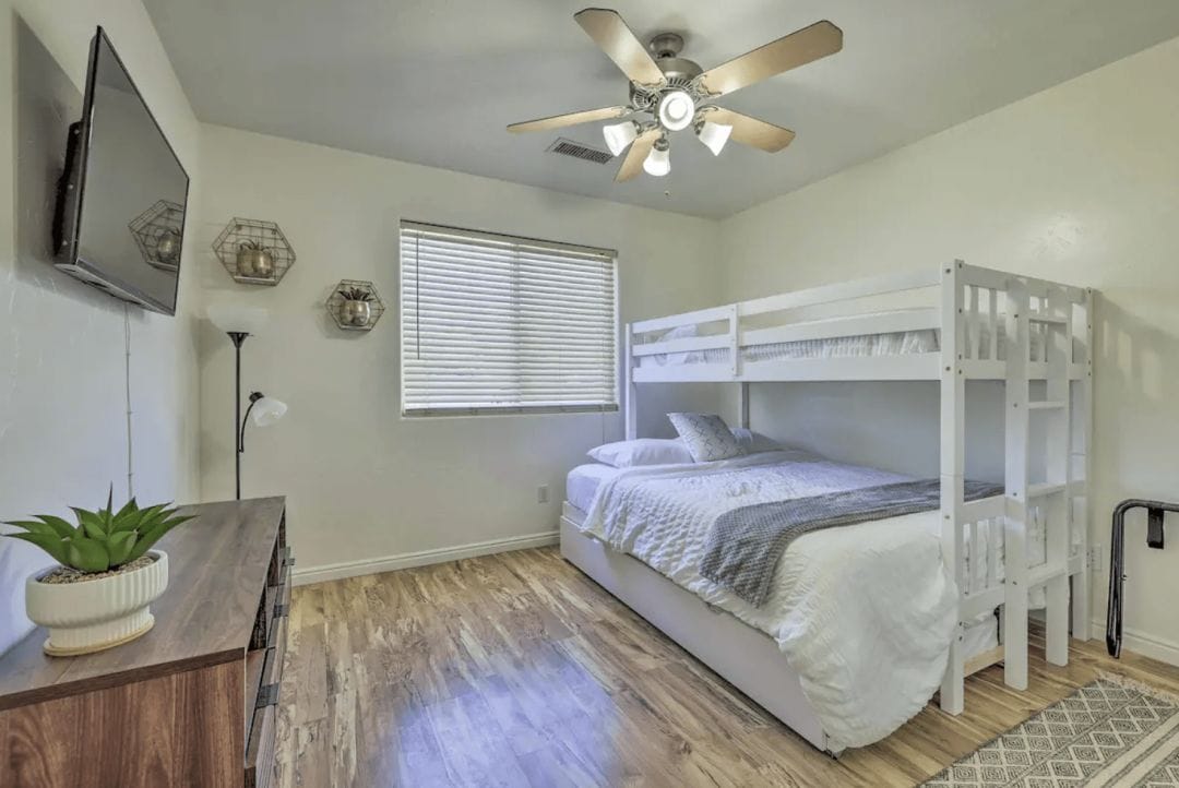 photograph of a white interior bedroom with a single bunkbed over a double bed with white and grey linens and a wood laminate floor, wall mounted television, wood dresser and overhead ceiling fan