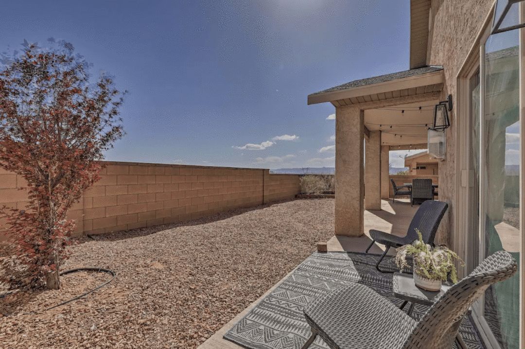 photograph of a xeriscaped side yard with 2 chairs and outdoor rug with a view of a patio at a single story house under a blue sky with white clouds in the distance