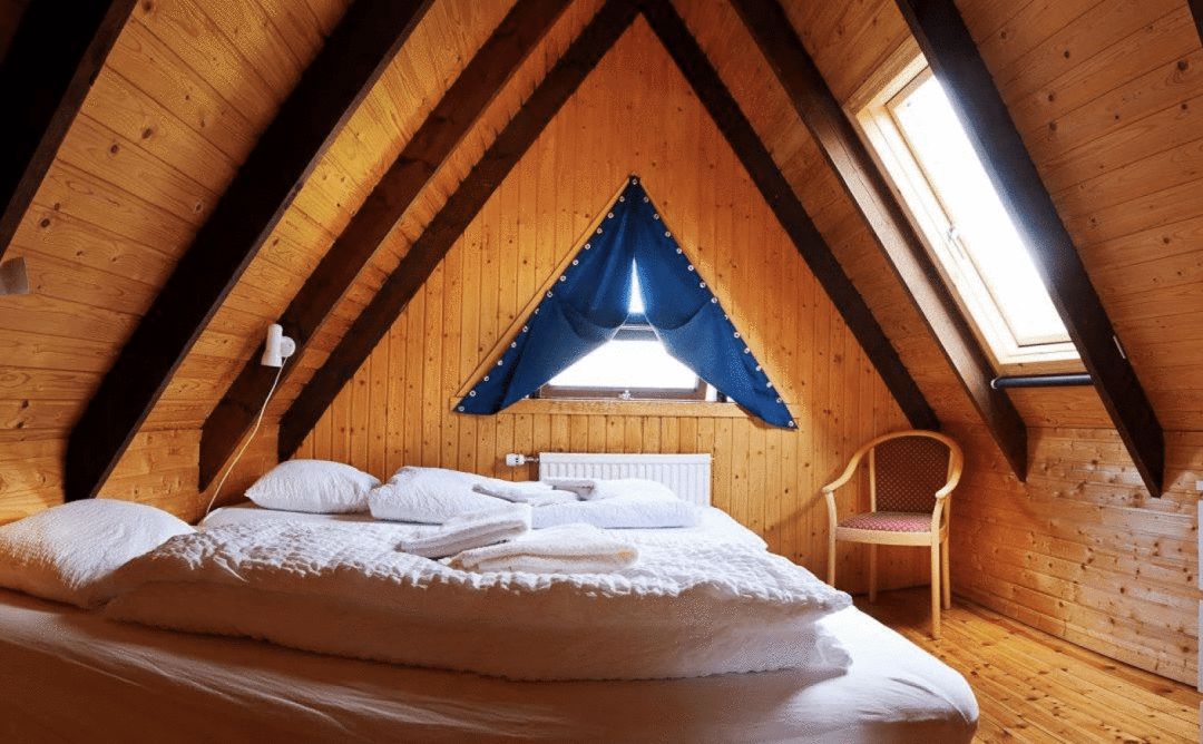 interior view of a bedroom with 2 single beds with white linens under a peaked vaulted wood ceiling with a window and blue curtains and a single wooden chair in the corner of the room