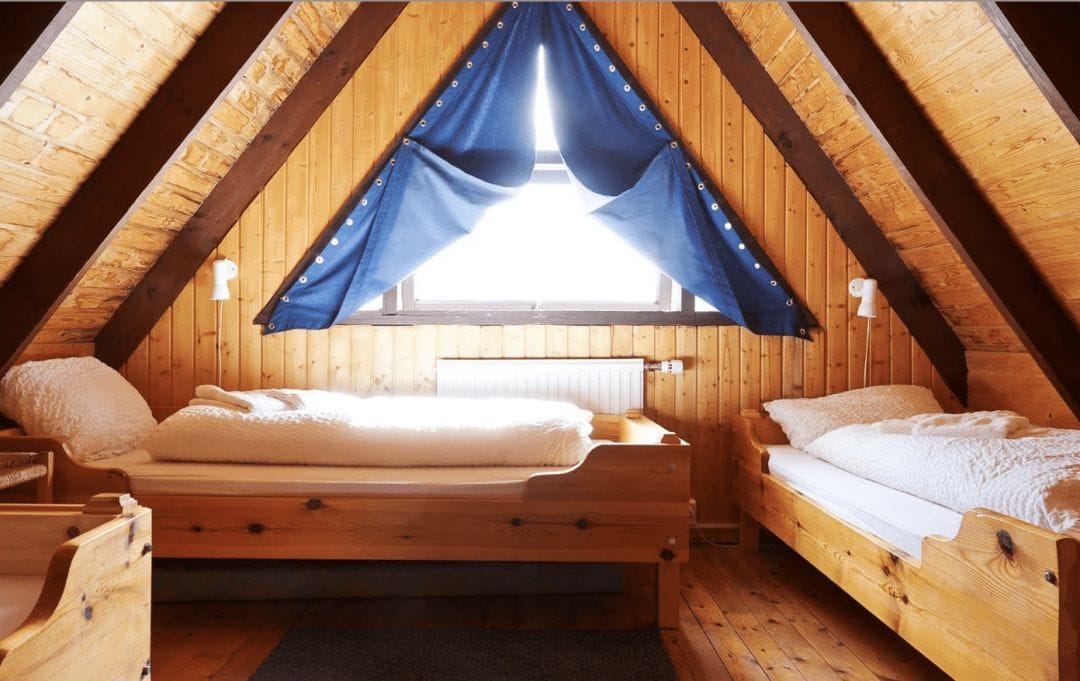 interior view of a bedroom with 3 single beds with white linens under a vaulted wood ceiling with a window and blue curtains