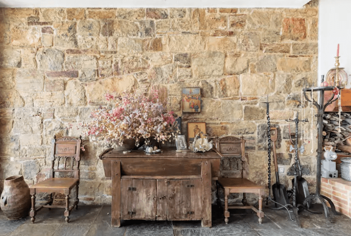 a photograph of an antique wood cupboard with dried flowers and 2 old wooden chairs in front of a large stone-faced wall