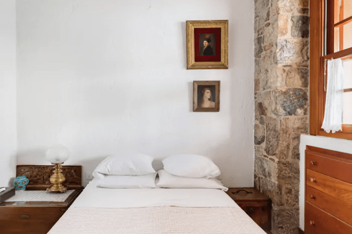 photograph of an bedroom interior in greece with a white wall with a large wooden cabinet and one large stone-faced wall with a window and a queen sized bed with white linens