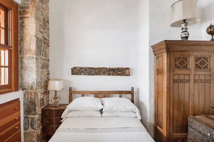 photograph of an bedroom interior in greece with a white wall with a large wooden cabinet and one large stone-faced wall with a window and a queen sized bed with white linens