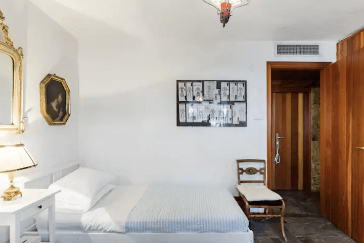 a bedroom in Greece with white walls a double bed a white nightstand and brown chair with a door open to a hallway with dark stone floors