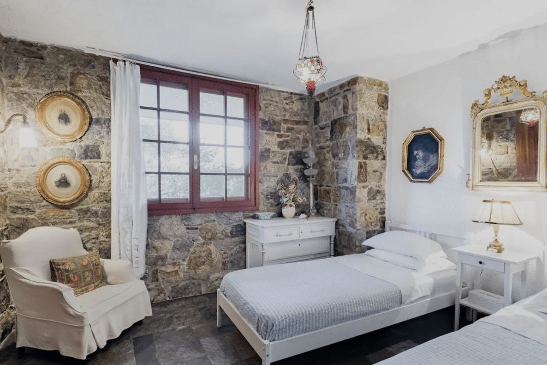 photograph of an bedroom interior in greece with a white wall with paintings one large stone-faced wall with window and 2 single beds a white nightstand a white side table and while chair