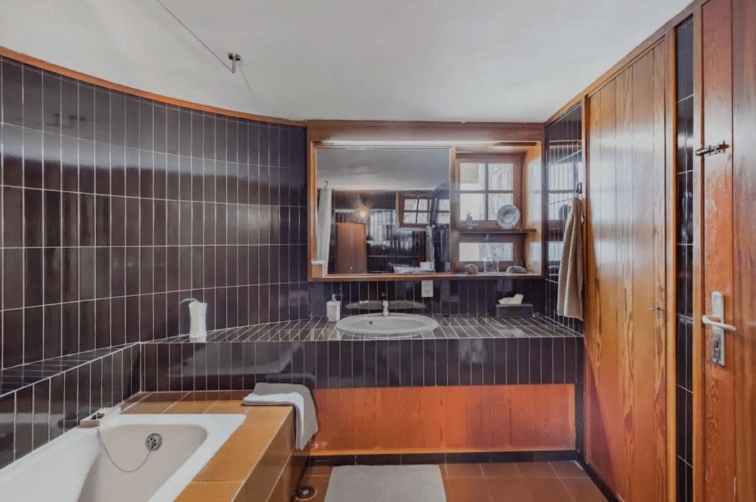 photograph of an interior bathroom in a house in Greece with black wall tile a white ceiling one white sink, one white tub and brown floor tiles