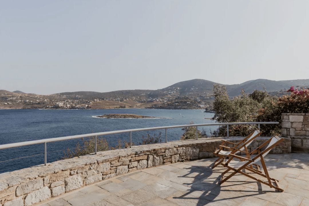 photograph of 2 empty white chairs on a stone patio overlooking an ocean and the coastal plains of Greece