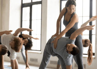 A woman with short brown hair wearing a grey top and grey bottoms stands above a male student in a studio of 5 yogis as they pose in extended side angle and the woman adjusts his pose