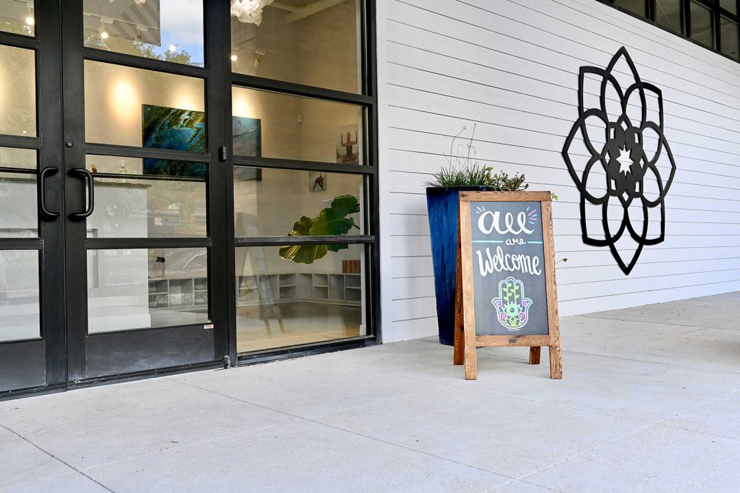 My Vinyasa Practice Donation Based Yoga Austin, TX Photo in front of a yoga studio, large windows, and planters, and mandala with a sign that says "all are welcome"