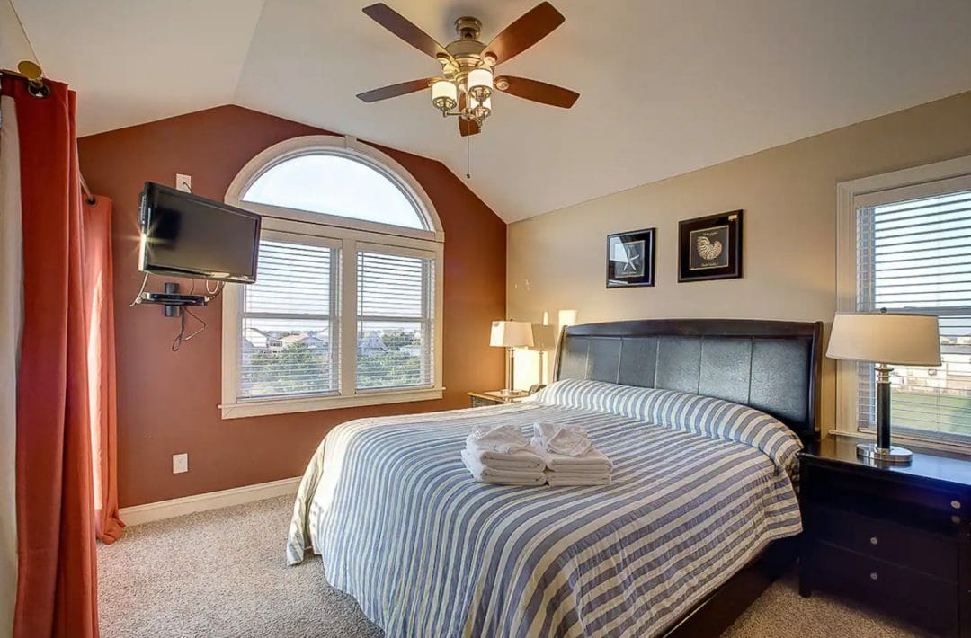 King sized bed with blue and white striped duvet, black leather headboard, red currtins, wall mounted TV, ceiling fan and large windows.
