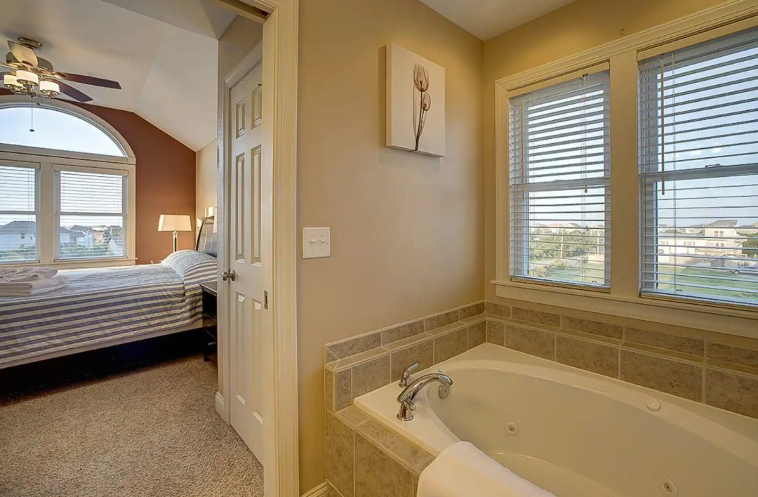 Tan bathroom with jetted soaker tub and view of bedroom with blue and white stripe duvet, ceiling fan and large window.