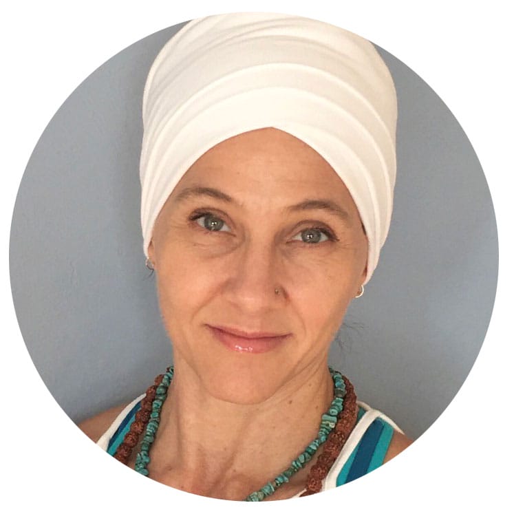 Headshot of Tina Hilbret: Woman with green eyes wearing white turban, wooden mala, and turquoise necklace