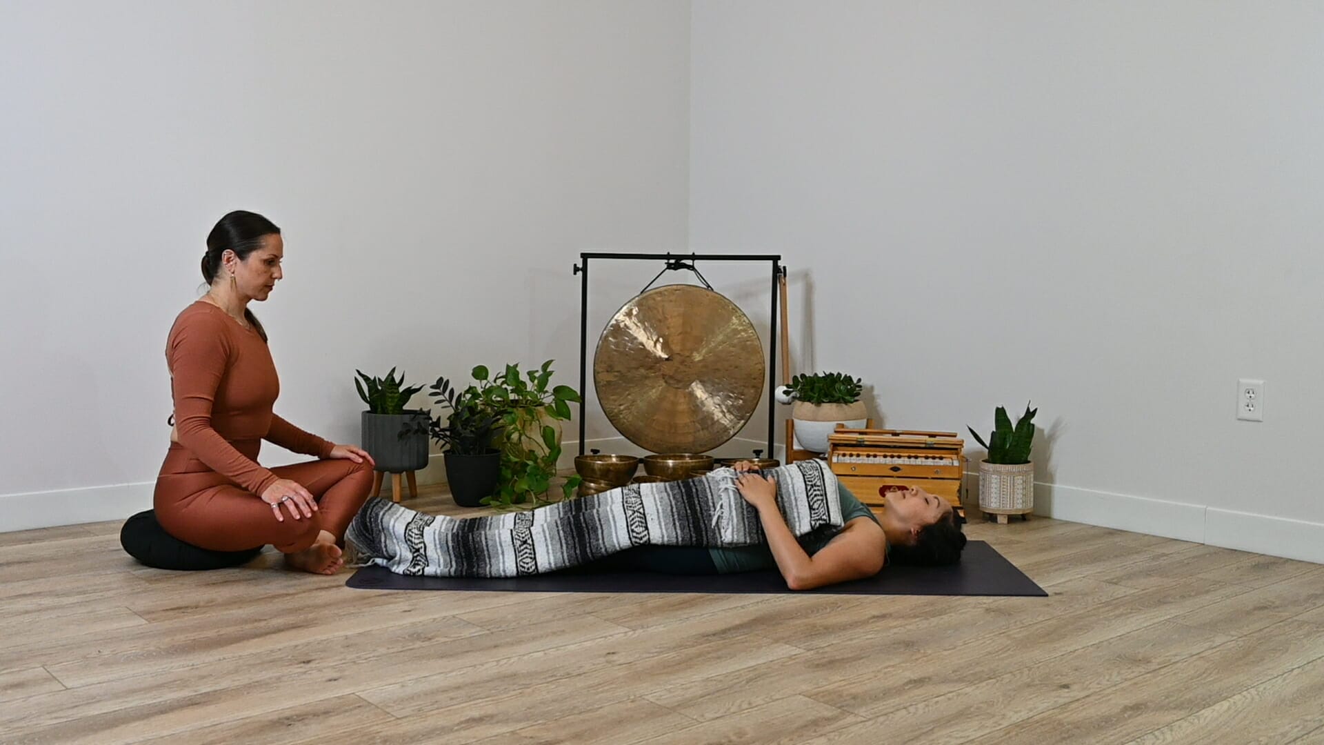 Student laying down with blanket over them while teacher sits at their feet reading yoga nidra.