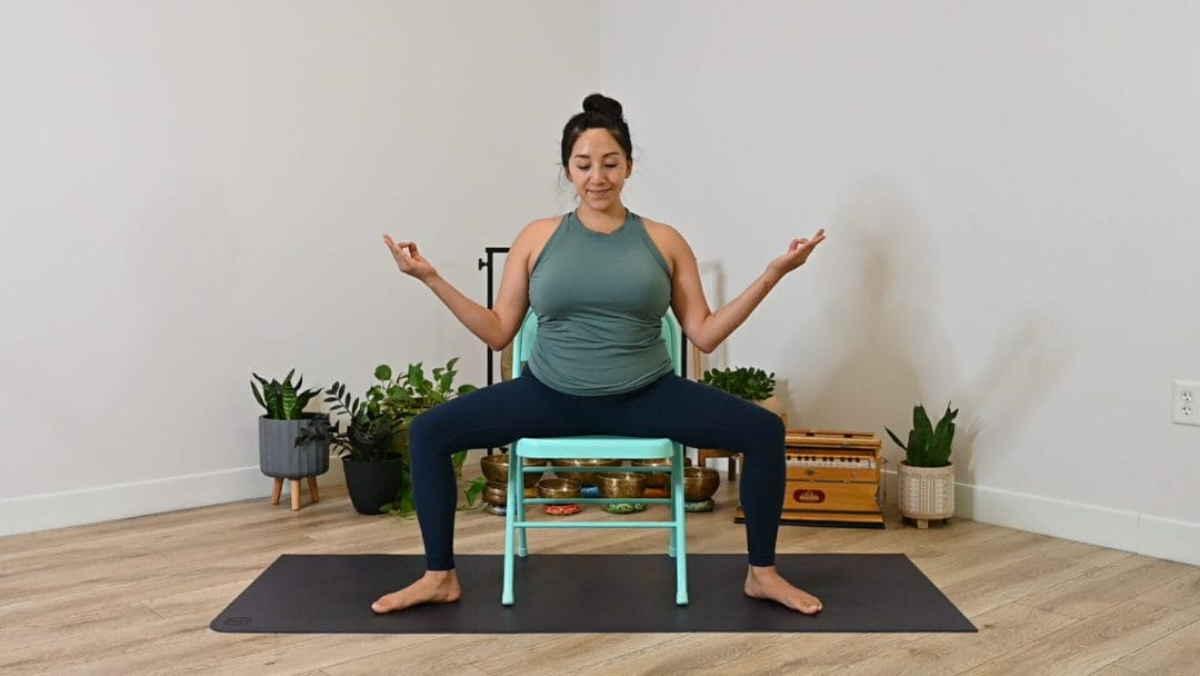 Hispanic woman practicing in seated goddess pose on a turquoise folding chair.