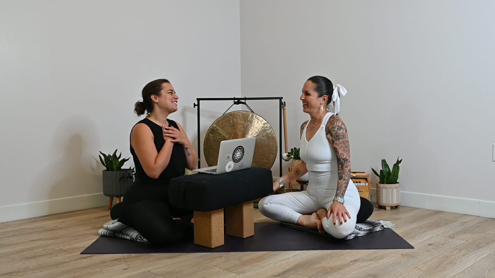 Tara Cleven and Michelle Young sit on a yoga mat in front of a laptop on blocks discussing coaching