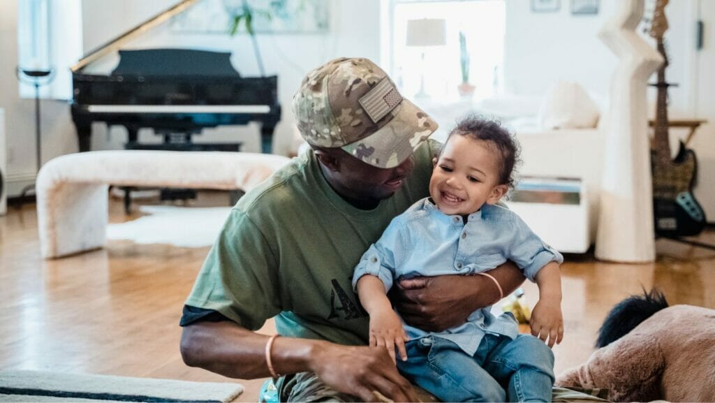 A man in a green shirt and green came hat with the American Flag emblem holds a young boy on his lap with black curly hair as they both smile.