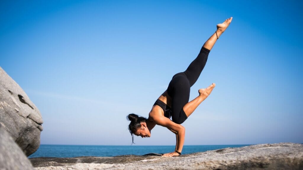 A woman with black hair in a bun does a variation of crow pose with one leg extended as she balances on a rock with the ocean in the background.