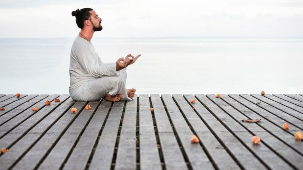A man dressed in all white with his brown hair in a bun sits on a wooden dock in meditation with Gyan mudra. 