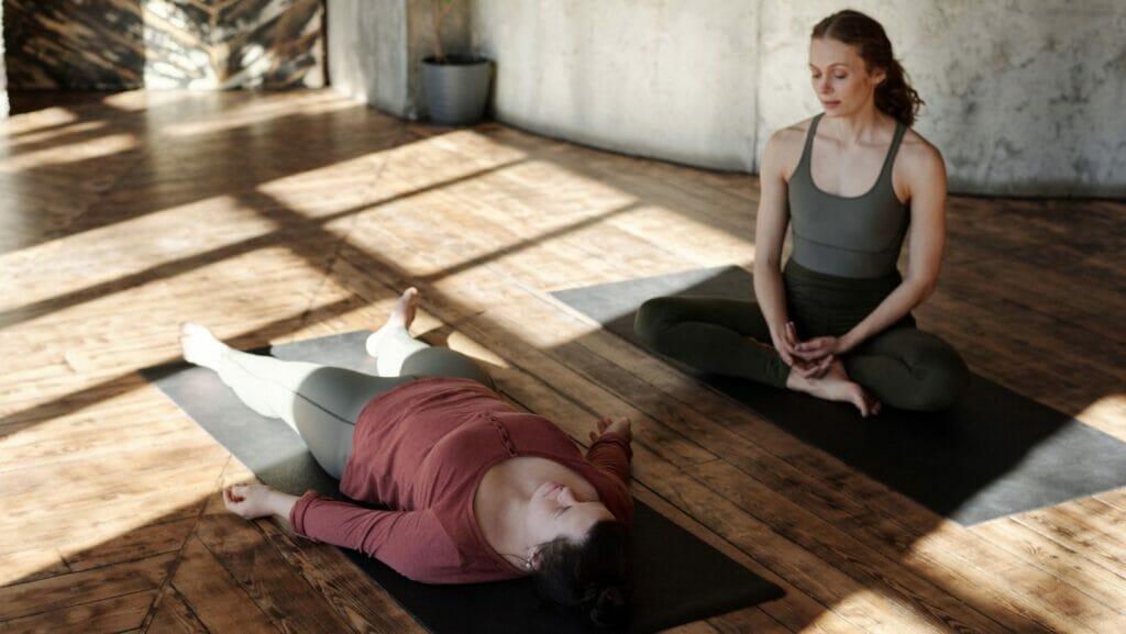 One woman in a dark green yoga outfit sits on a black yoga mat and gazes at another woman in a red shirt and gray pants lying on a black mat in rest.