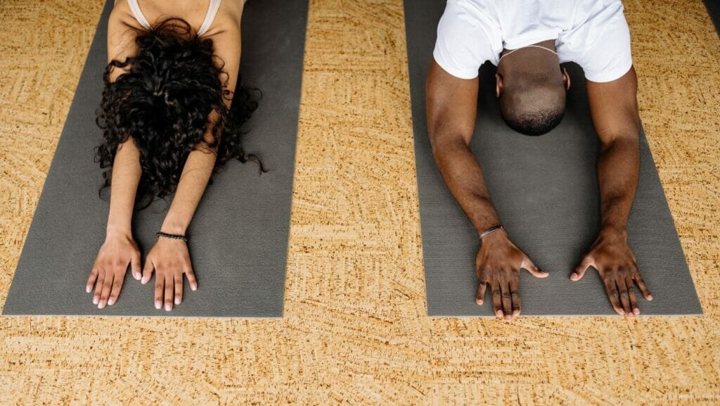 Two people lie on black yoga mats side by side in child's pose.