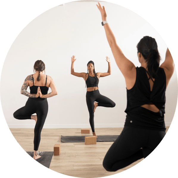 3 women yogis in a yoga class. They are balanced on 1 foot. The other leg is bent with the foot resting on the opposite leg. Their hands are raised in the air.