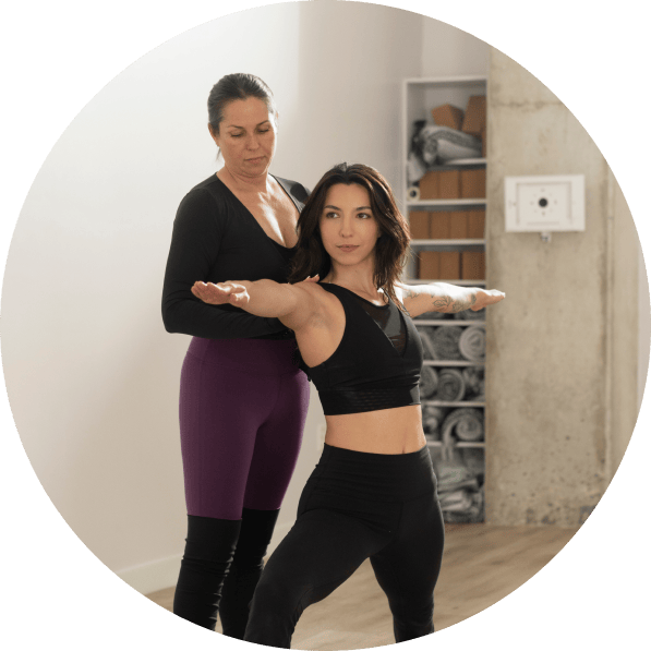 Two women yogis are standing in a warrior 2 yoga pose. One women is assisting the other with proper form.