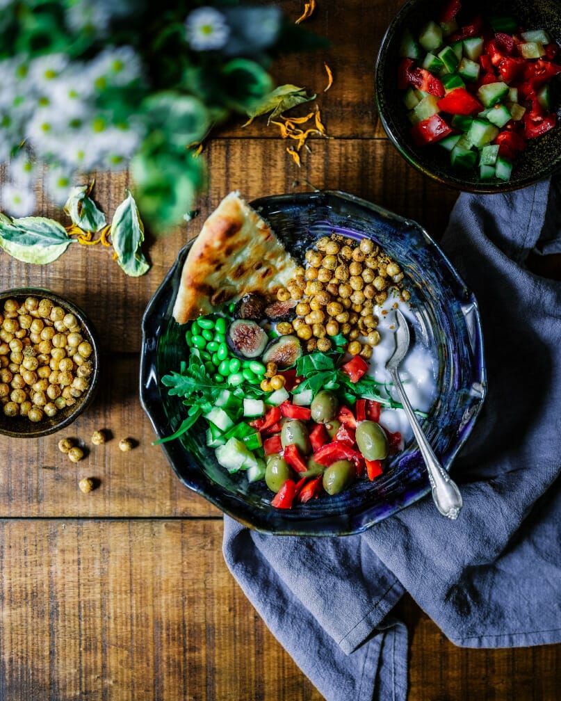 A bowl of food sits on a wooden table. The food contains vibrant colors of beans, peppers, and vegetables.