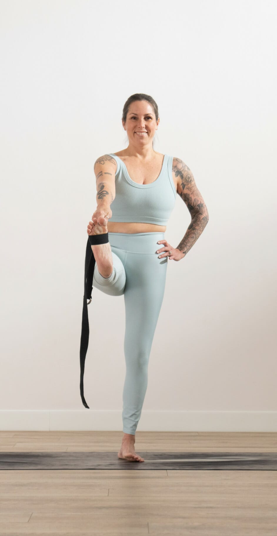 A photo of Michelle, standing on a yoga mat with her left leg held straight out in front of her. She is using a band to hold her foot, and is smiling with her left hand on her hip.