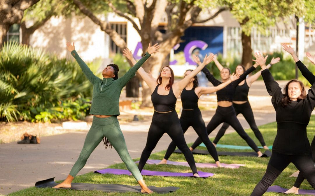 A yoga class outside on the grass. The women are standing with both arms up above their heads.