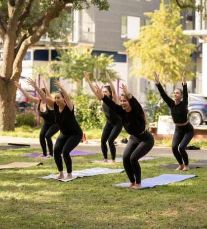 A group of women yogis are outside the MVP studio doing yoga on the lawn. Their knees are bent, and their arms are raised above their heads.