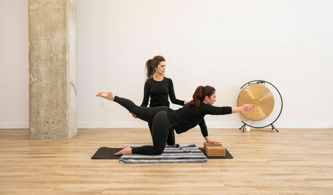 Two women yogis are practicing yoga in the MVP studio. One woman assists the other with proper form in a yoga pose.